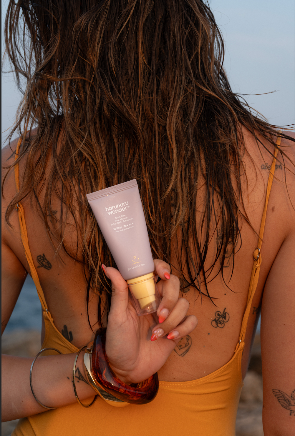 Mannequin à la plage montrant la crème solaire pure mineral relief riz noir Haruharu Wonder dans son dos, maillot de bain et tatouage visibles