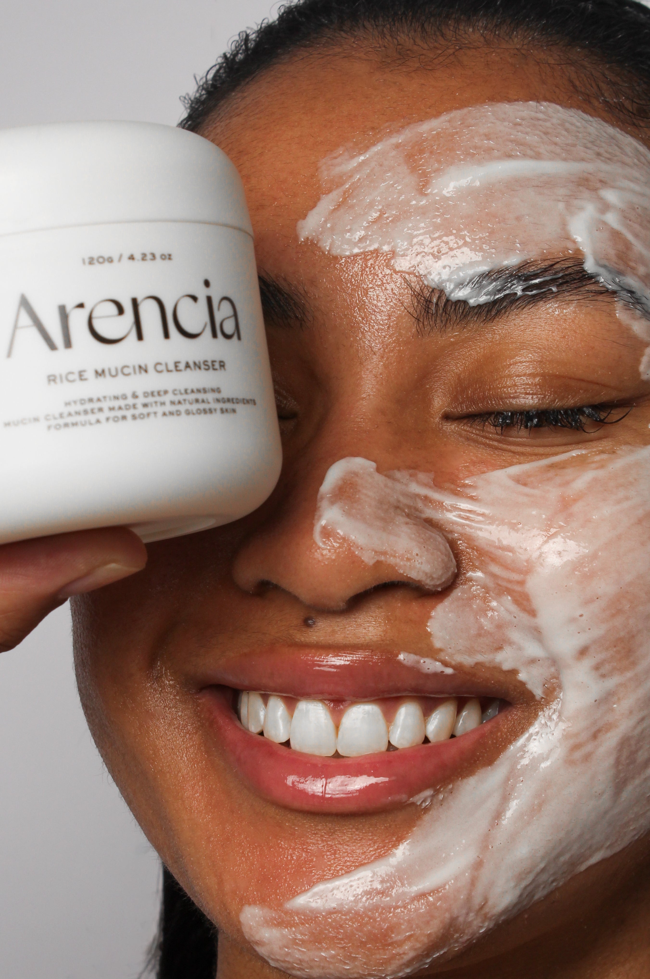Mannequin avec le nettoyant à la mucine de riz Arencia appliqué sur le visage, tenant le pot du produit